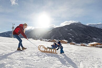 Eine Mutter zieht im Winter einen Schlitten, auf dem zwei kleine Kinder sitzen direkt hinterm Almfamilyhotel Scherer in Tirol 