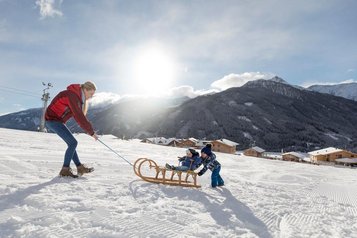 Eine Mutter zieht im Winter einen Schlitten, auf dem zwei kleine Kinder sitzen direkt hinterm Almfamilyhotel Scherer in Tirol 