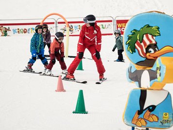 Skikurs für Kinder: Skilehrer in rotem Anzug übt mit drei Kindern zwischen Slalomkegeln, im Vordergrund eine große Pinguin-Figur.
