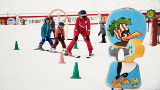 Skikurs für Kinder: Skilehrer in rotem Anzug übt mit drei Kindern zwischen Slalomkegeln, im Vordergrund eine große Pinguin-Figur.