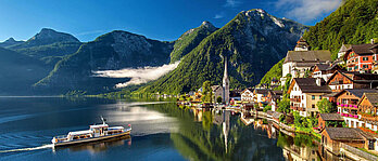 Ein Schiff fährt über einen See zu einer Anlegestelle an einem Ort. Im Hintergrund Bergkulisse der Alpen.