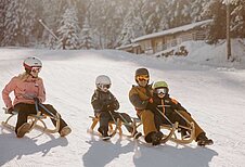 Eine Familie hat Spaß beim Rodeln auf einer sonnenbeschienenen Piste im Winter am Hotel Habachklause im Salzburger Land, umgeben von malerischen Schneelandschaften.