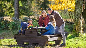 Eine Gastfamilie vom Familienhotel Am Rennsteig sitzt im Thüringer Wald auf einer Sitzbank und legen eine Pause ein.