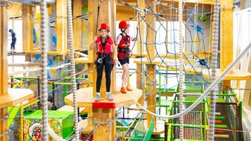 Zwei Kinder in Sicherheitsausrüstung gehen über Holzplattformen in einem bunten Indoor-Hochseilgarten im Hotel Feldberger Hof, umgeben von Seilen und Netzen.