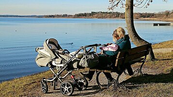 Frau sitzt auf einer Bank am Seeufer und hält ihr Baby im Arm. Daneben steht der Kinderwagen.