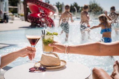 Zwei Cocktails am Beckenrand, im Hintergrund spielende Kinder im Wasser und eine rote Wasserrutsche.