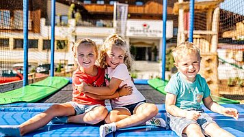 Drei Kinder sitzen auf dem Trampolin auf dem Außengelände des Familienhotels Das Hopfgarten.