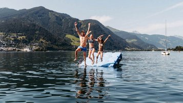 Kinder springen gemeinsam von einer Badeplattform in den Zeller See direkt beim Familienhotel Amiamo, mit Bergpanorama im Hintergrund.