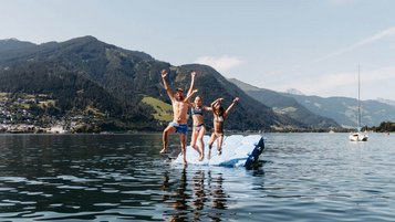 Kinder springen gemeinsam von einer Badeplattform in den Zeller See direkt beim Familienhotel Amiamo, mit Bergpanorama im Hintergrund.