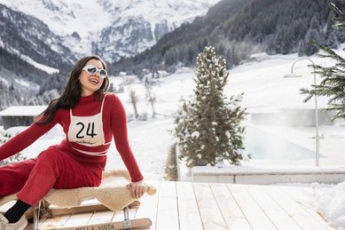 Frau sitzt im Winter im Hotel Bella Vista in Südtirol auf einem Schlitten im Schnee und lächelt, im Hintergrund eine verschneite Berglandschaft.