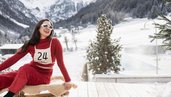 Frau sitzt im Winter im Hotel Bella Vista in Südtirol auf einem Schlitten im Schnee und lächelt, im Hintergrund eine verschneite Berglandschaft.