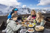 Eine Familie sitzt bei einer Mahlzeit vor einer Almhütte im Winterurlaub am Familienhotel Gorfion in Liechtenstein.