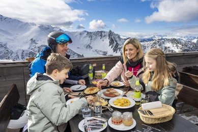 Eine Familie sitzt bei einer Mahlzeit vor einer Almhütte im Winterurlaub am Familienhotel Gorfion in Liechtenstein.