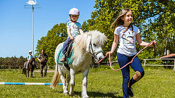 Ein Mädchen sitzt auf einem Pony und wird von einem anderen Mädchen über Hindernisse geführt.