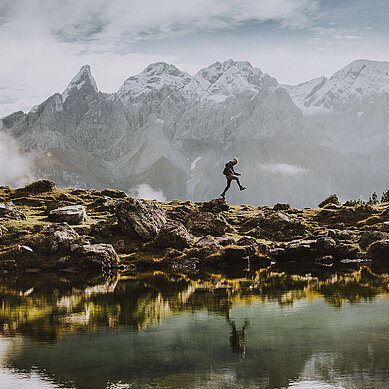 Ein Mann springt und wandert über Felsen. Davor ist ein See und dahinter ist ein Gebirge im Nebel im Allgäu.