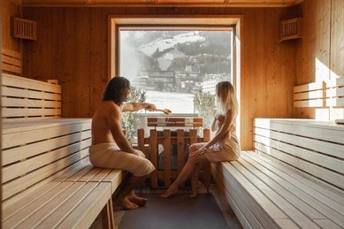 Eltern sitzen in der Sauna des Familienhotel Mia Alpina in Tirol mit Holzwänden, großem Fenster und winterlicher Bergkulisse.