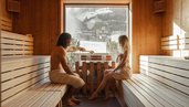 Eltern sitzen in der Sauna des Familienhotel Mia Alpina in Tirol mit Holzwänden, großem Fenster und winterlicher Bergkulisse.