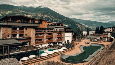 Außenansicht des Familienhotel Mia Alpina in Tirol mit Pool, Liegen, Sonnenschirmen und Bergen im Hintergrund.