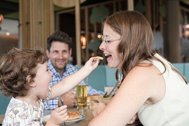 Ein kleines Mädchen füttert lachend seine Mama mit einer Pommes im Familienhotel beim Mittagessen.