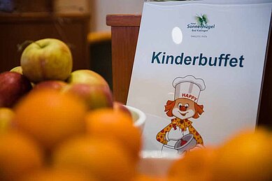 Ein Schild für das Kinderbuffet mit dem Logo des Familienhotels Sonnenhügel und einer fröhlichen Clownfigur, umgeben von einer Auswahl an frischem Obst im Vordergrund.