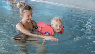 Eine Frau unterstützt ein Kind mit Schwimmhilfe beim Schwimmen im Hallenbad des Familienhotels Gorfion in Liechtenstein.