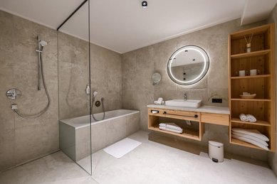 Großzügiges Badezimmer im Hotel Zauchenseehof mit Badewanne, begehbarer Glasdusche, hellen Natursteinfliesen, rundem beleuchtetem Spiegel über dem Waschtisch und offenem Holzregal mit Handtüchern und Kosmetikartikeln.