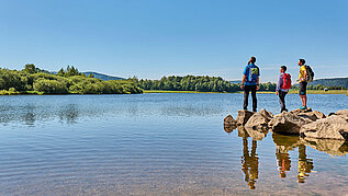 Drei Wandere stehen am Perlsee im Bayerischen Wald und genießen die Sonne.