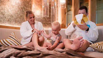 Ein Vater im Bademantel pustet einen Ballon auf, die Mutter sitzt lächelnd daneben und hält die Hand ihres kleinen Kindes, das auf einer Couch im gemütlichen Wellnessbereich des Familienhotels Huber in Südtirol sitzt.