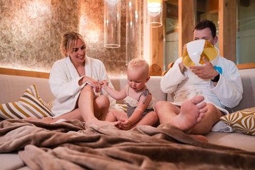 Ein Vater im Bademantel pustet einen Ballon auf, die Mutter sitzt lächelnd daneben und hält die Hand ihres kleinen Kindes, das auf einer Couch im gemütlichen Wellnessbereich des Familienhotels Huber in Südtirol sitzt.