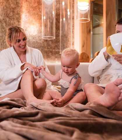 Ein Vater im Bademantel pustet einen Ballon auf, die Mutter sitzt lächelnd daneben und hält die Hand ihres kleinen Kindes, das auf einer Couch im gemütlichen Wellnessbereich des Familienhotels Huber in Südtirol sitzt.