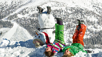 Zwei Kinder liegen im Schnee neben einem Schneemann und strecken die Beine in die Höhe.