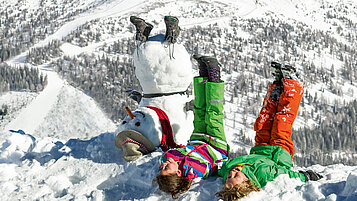 Zwei Kinder liegen im Schnee neben einem Schneemann und strecken die Beine in die Höhe.