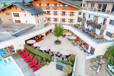 Luftaufnahme des Familienhotels Amiamo mit hellen Fassaden, roten Sonnenliegen am Außenpool, beschatteter Terrasse mit Tischen und Schirmen sowie angrenzendem Spielbereich.