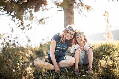 Zwei Mädchen, die sich im Rahmen der Kinderbetreuung als Indianer verkleidet haben, sitzen auf der grünen Wiese unter zwei Bäumen. Im Family Home Alpenhof in Südtirol wird eine liebevolle Baby- und Kinderbetreuung angeboten.