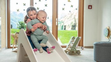 Mädchen und Baby sitzen gemeinsam auf einer weißen Rutsche in einem hellen Spielzimmer.