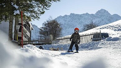 Kind mit Helm und Skiausrüstung steht auf verschneiter Piste vor einer malerischen Bergkulisse, was Wintersportaktivitäten für Kinder in einem familienfreundlichen Hotelumfeld veranschaulicht.