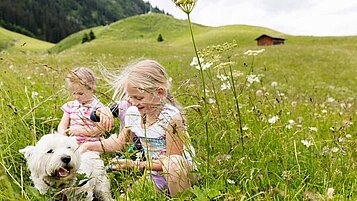 Zwei Mädchen spielen beim Wandern im Familienurlaub in Österreich mit ihrem Hund in einer schönen grünen Wiese.