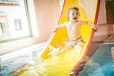 Kleiner Junge rutscht im Kinderbecken die Wasserrutsche im Schwimmbad viel Freude hinunter im Familienhotel Landgut Furtherwirt in Tirol.
