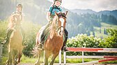 Zwei Kinder reiten auf zwei Pferden auf dem Reitplatz des Familienhotels Landgut Furtherwirt. Auf dem Reitplatz vom Landgut Furtherwirt gibt es jeden Tag die Möglichkeit für einen Ausritt oder eine Reitstunden. 
