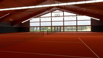 Innenansicht der modernen Tennishalle im Familienhotel Sonnenhügel mit hell erleuchteten, roten Tennisplätzen und großen Fenstern mit Ausblick auf die grüne Landschaft.