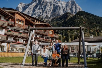 Hotelteam sitzt und steht an einer Schaukel vor einem Alpenhotel mit markanter Bergkulisse.