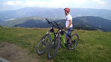 Ein Junge steht in mit zwei E-Mountainbikes auf einer grünen Wiese. Im Hintergrund ist die schöne Kärntner Berglandschaft zu sehen.