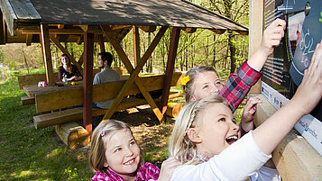 Kinder entdecken eine Station auf dem Walderlebnispfad im Füllener Wald in Meppen. Die Erwachsenen sitzen an einem überdachten Picknickplatz. 