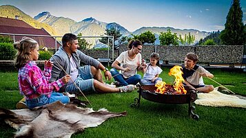 Eine Familie sitzt im Garten vom Adler Familien- & Wohlfühlhotel in Tirol und braten Marshmallows am Lagerfeuer.