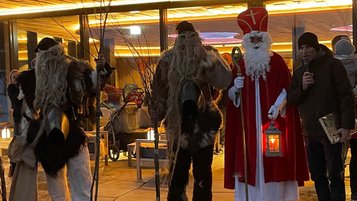Nikolaus und zwei als wilde Gestalten verkleidete Begleiter stehen gemeinsam mit dem Sprecher Christian Neusch vor dem Eingang eines beleuchteten Familienhotels während einer adventlichen Veranstaltung.