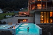 Neubau des Familienhotel Bella Vista in Südtirol bei Nacht mit beleuchtetem Infinity Pool und Blick zur Kirche.