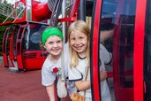 Zwei fröhliche Kinder in roten Gondel-Spielgeräten auf dem Outdoor-Spielplatz des Hotel Engel im Schwarzwald