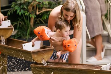 Mutter hält ihr Kleinkind mit Schwimmflügeln über einen Wasserspielbereich im Zauchenseehof, während zwei Erwachsene im Bademantel lächelnd von der Treppe aus zusehen.
