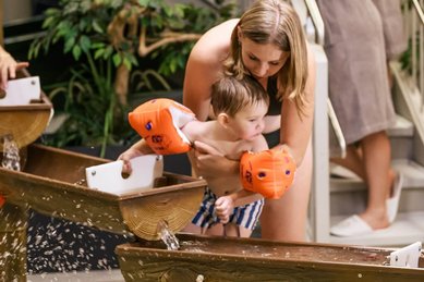 Mutter hält ihr Kleinkind mit Schwimmflügeln über einen Wasserspielbereich im Zauchenseehof, während zwei Erwachsene im Bademantel lächelnd von der Treppe aus zusehen.