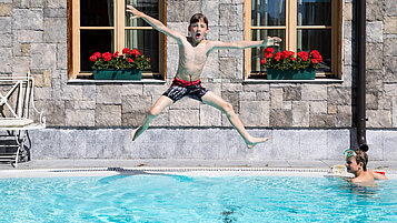 Jugendlicher Junge springt lachend in den Außenpool des Familienhotels Tirolerhof in Tirol.